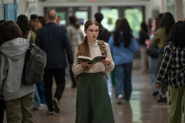 Teen girl with a backpack reads a book while standing in a crowded school hallway with students walking around her.