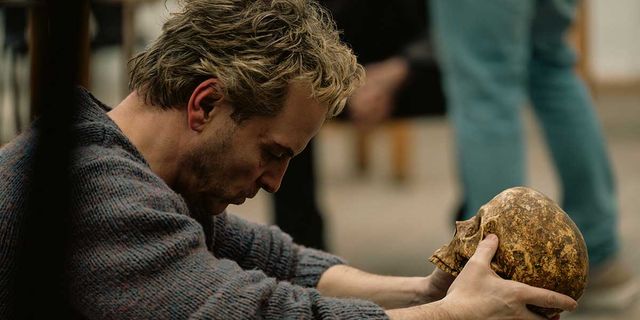 A man sits indoors leaning forward while holding a weathered human skull, with blurred standing figures behind him.