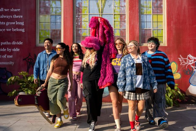 Seven people in colourful clothing walk along a sunny pavement in front of a red building.