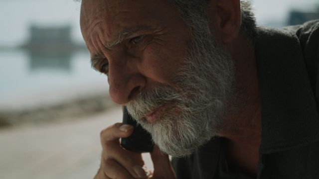 A grey-bearded man holds a phone to his ear outdoors.