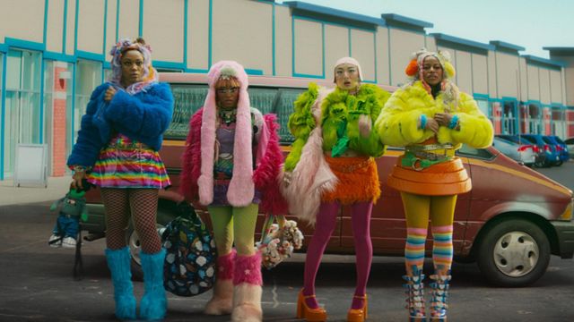 Four people in bright furry colourful outfits stand in a parking lot in front of a red minivan and strip-mall building.
