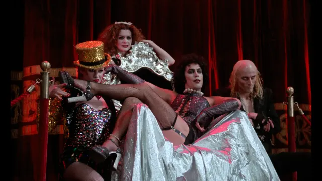 Four elaborately costumed performers pose on an ornate silver throne before red stage curtains, with sequins, fishnets and dramatic cabaret-style makeup.