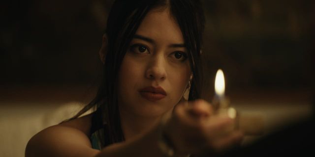 A woman holds a small flame in a dim indoor setting, light falling across her face.