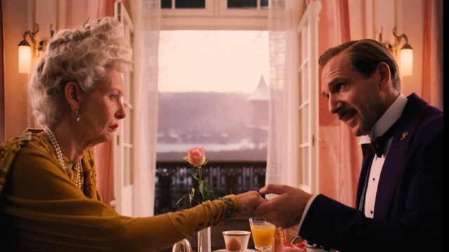 An older woman and a man in formal evening wear sit at a restaurant table by open balcony doors, holding hands beside a pink rose and breakfast dishes.