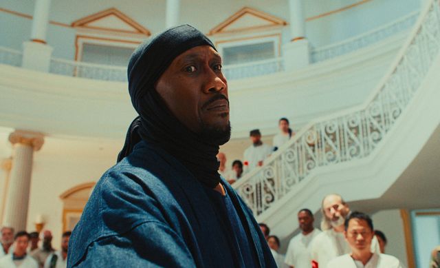 A serious-looking man in a dark blue head covering and robe stands in the foreground of a grand white interior with ornate staircases and people in white gathered behind him.
