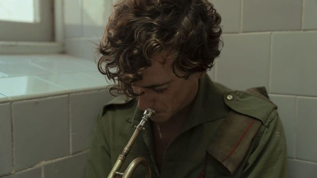 A curly-haired man in a green shirt plays a brass mouthpiece in a tiled room.
