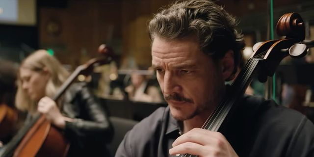Close-up of a serious male cellist in a dark suit playing in an orchestra, with another blurred cellist and music stands in the background.