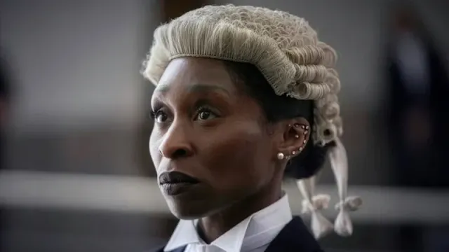 Close-up of Cynthia Erivo in a barrister’s wig and dark court attire, with a courtroom blurred behind her.