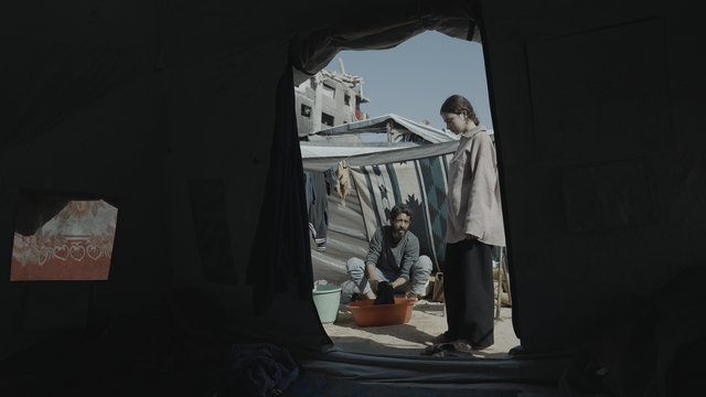 Inside a dark tent, a young person stands beside a crouching man washing clothes in a basin, with tarps and a damaged building behind them.