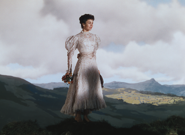 Woman in a long pale dress holding flowers on a grassy ridge, with cloudy sky and distant mountains.