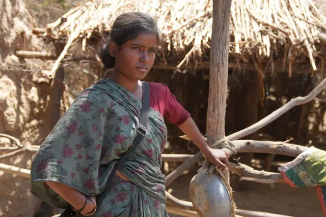 A young woman in a floral green sari stands beside a wooden fence in front of a thatched rural structure, holding a metal pot.