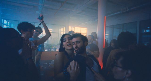 A crowd dances in a smoky indoor venue lit in blue, beside a vertical red neon strip.