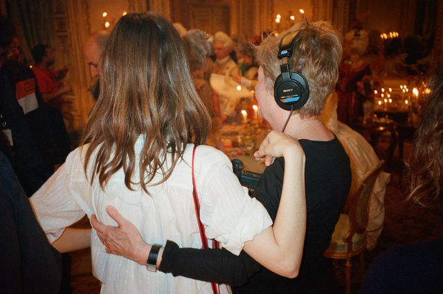 Two people seen from behind with arms around each other in a crowded, candlelit ornate room; one wears large headphones.