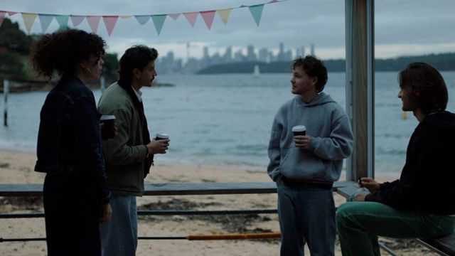 Four people holding takeaway coffee cups talk under bunting in a beachfront shelter, with water and a distant city skyline behind them.