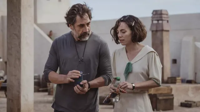 A man and woman walk across a rooftop set holding drinks and sunglasses.