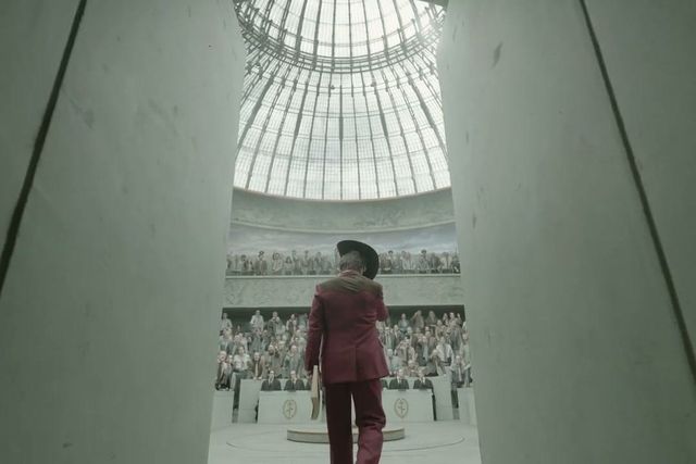 Person in a burgundy suit and black hat walks into a large domed auditorium filled with seated spectators.