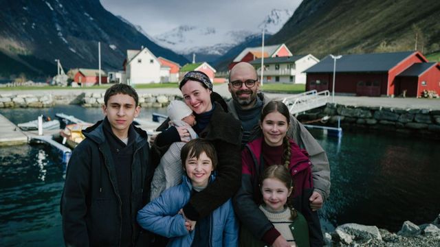 Two adults and four children, including a baby held by one adult, pose beside dark water and small boats in a mountain village with red, white and green buildings under snow-capped peaks.