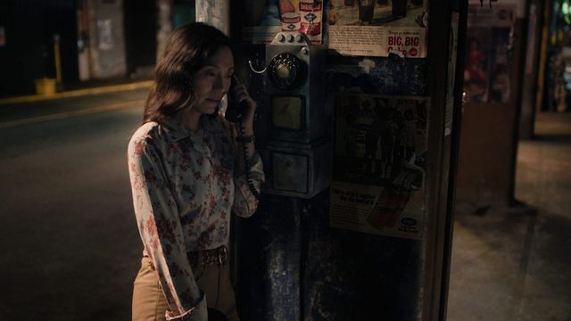 A woman in a white blouse with red floral prints and tan trousers uses a black rotary payphone in a dim, warm-toned street-side booth covered with faded advertisements.