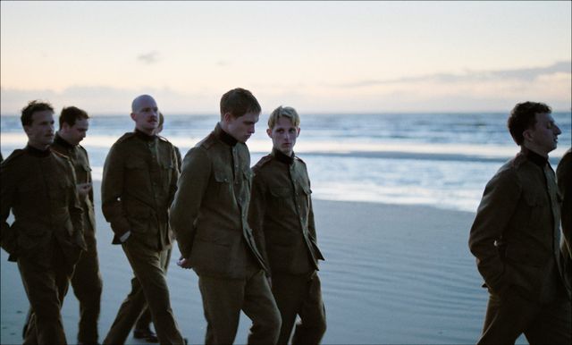 Six uniformed men walk along a beach at dusk with ocean waves in the background.
