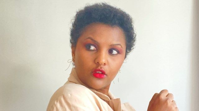 Woman with short curly black hair, red lipstick, reddish eyeshadow, hoop earrings, and beige top, glancing sideways.