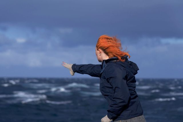 Red-haired woman in dark jacket and knit gloves reaching toward a stormy sea, wind blowing her hair, overcast sky.