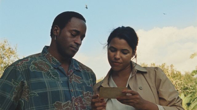 Two people standing outdoors looking at a small card or photograph the woman is holding, with blue sky and greenery behind them.