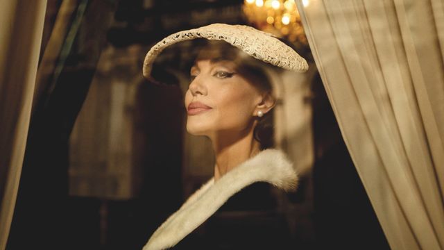 Elegant woman in a wide-brimmed white hat, pearl earring, and fur-trimmed coat, framed by curtains in warm lighting.