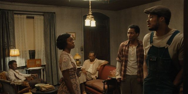 Five people in a dimly lit 1950s living room with vintage furniture and radio, appearing tense during a serious conversation.