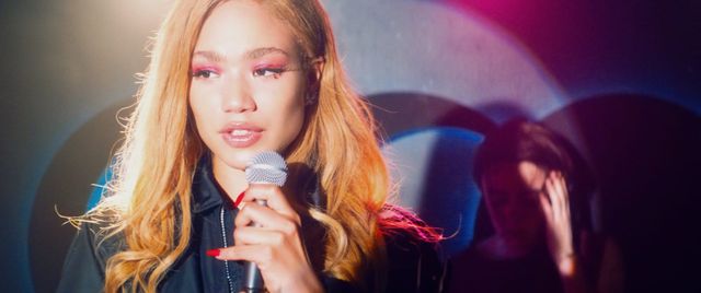 Young woman with blonde hair and pink eyeshadow holding a microphone, wearing a black jacket with red nails, stage lighting behind her.