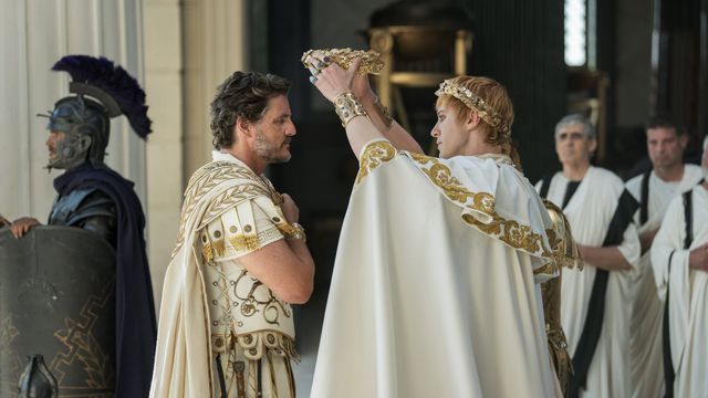 Young man in white Roman emperor robes places a golden crown on a kneeling man in Roman attire, with guards and senators behind.