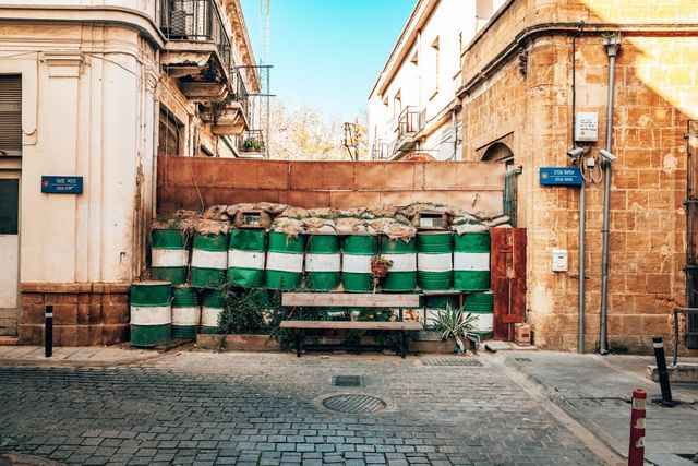 Green-and-white oil drum barricade topped with sandbags blocking a street in Nicosia's UN Buffer Zone, Cyprus, with street signs and a bench.