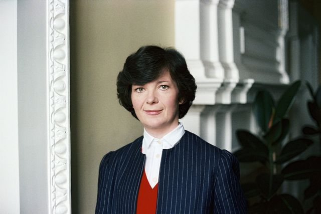Woman with short dark hair wearing a navy pinstripe blazer, red sweater, and white collared shirt, standing by ornate white molding.