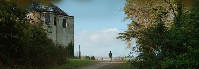 Lone figure walks on a path beside an abandoned building with a damaged roof, flanked by lush green trees under cloudy sky.