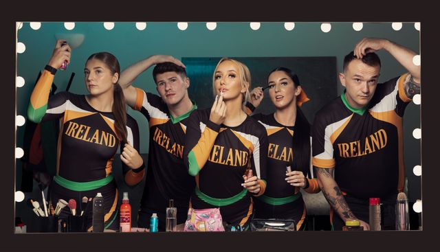Five Irish cheerleaders in matching black, orange, and green "IRELAND" uniforms getting ready at a lit vanity mirror backstage.