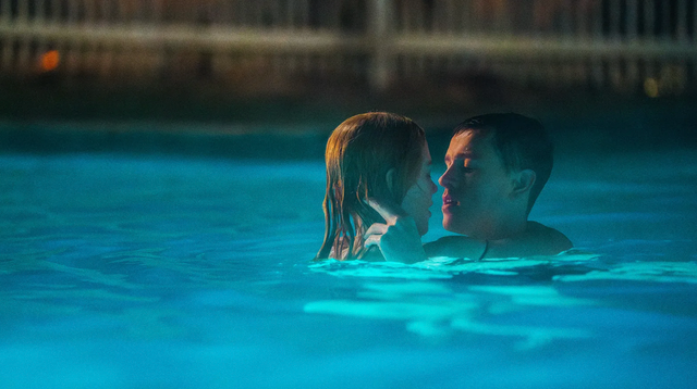 Two people embracing closely in a blue-lit swimming pool at night, faces near each other in an intimate moment.