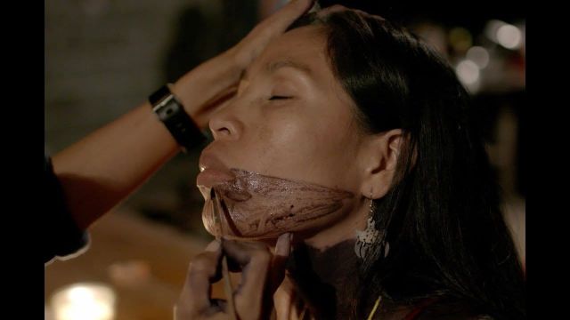 Makeup artist applying dark prosthetic or special effects makeup to a woman's chin and jawline, her eyes closed, dark hair down.