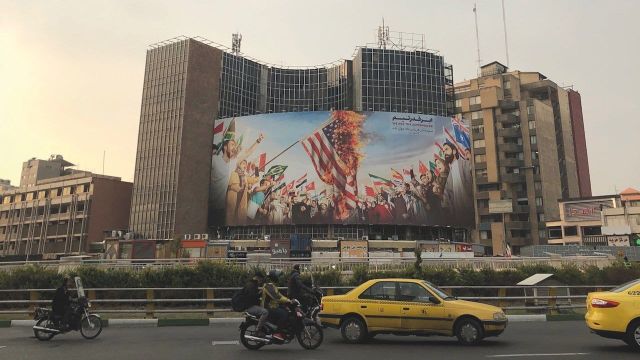 Large anti-American billboard in Tehran showing crowds burning U.S. and UK flags, with Persian text, motorcycles and yellow taxis on street below.