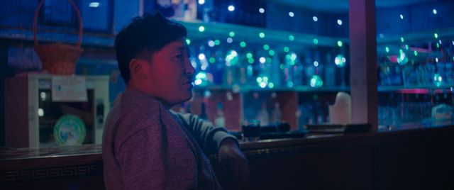 Young Asian man sitting at a dimly lit bar counter, illuminated by blue and green bokeh lights, looking to the right.