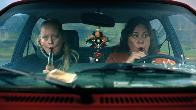 Two women sitting in a red car, each sipping from bottles through straws, with a decorative ornament hanging from the rearview mirror.