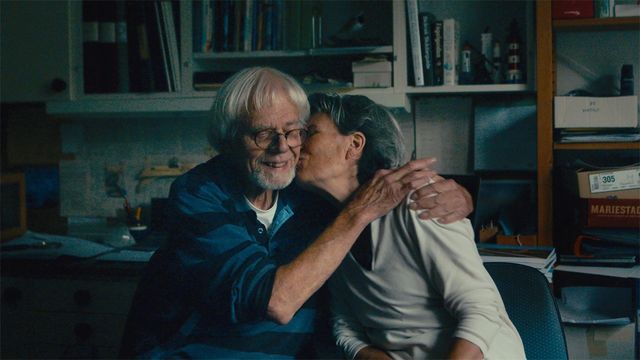 Elderly couple embracing in a cluttered home office; woman kisses smiling man with white hair and glasses on the cheek.