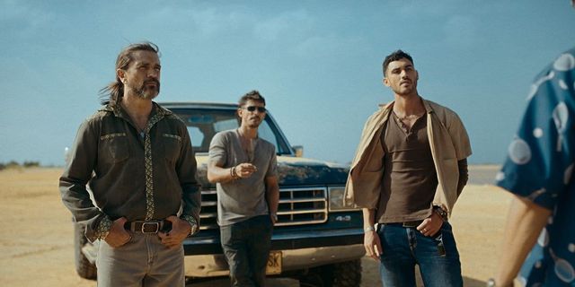 Three men stand in a desert landscape near a dark blue pickup truck, wearing rugged casual clothing, looking off-camera.