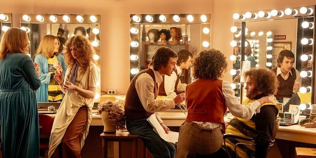 Backstage dressing room scene with actors in 1970s costumes and wigs near lit vanity mirrors with Hollywood-style bulbs.