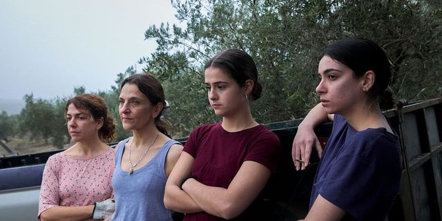 Four women of varying ages stand outdoors near olive trees and a truck, looking somber with arms crossed or resting.
