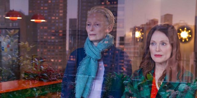 Two people standing by a window with city buildings reflected in the glass, one wearing a teal scarf, the other in a green patterned jacket.
