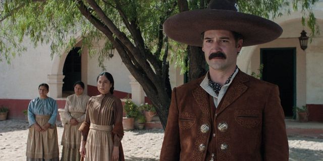 Man in brown embroidered charro suit and wide-brimmed hat stands in a courtyard, three women in period dress behind him.