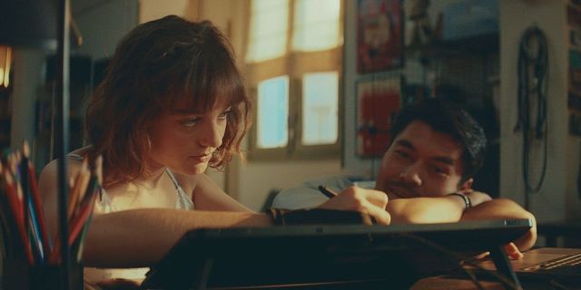 Young woman drawing on a digital tablet while a man rests his chin on his arms beside her, warmly lit creative workspace.