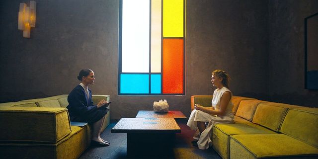 Two women sit facing each other on yellow sofas in a dark room with a colorful Mondrian-style stained glass window behind them.