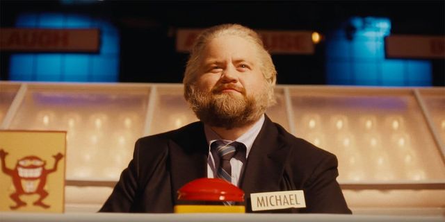 Bearded man in dark suit with striped tie sits at game show contestant desk with name placard "MICHAEL" and red buzzer.