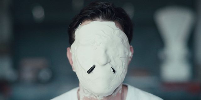 Person wearing a smooth white featureless mask covering their entire face, with two small dark slits near the nose area.