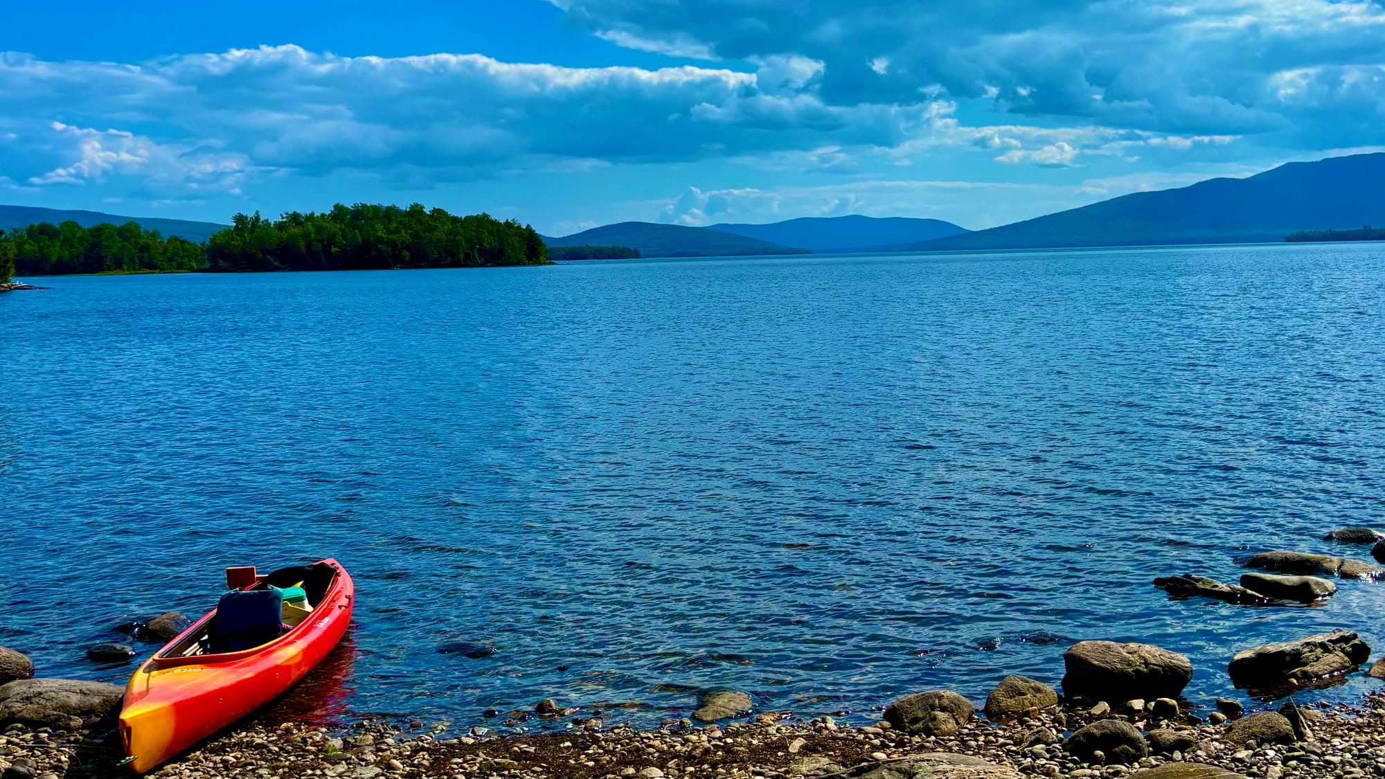 A red kayak resting on a pebbled shoreline with a scenic view of calm blue waters, distant mountains, and a partly cloudy sky.